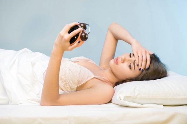 Eine Frau liegt im Bett und schaut verzweifelt auf ihren Wecker